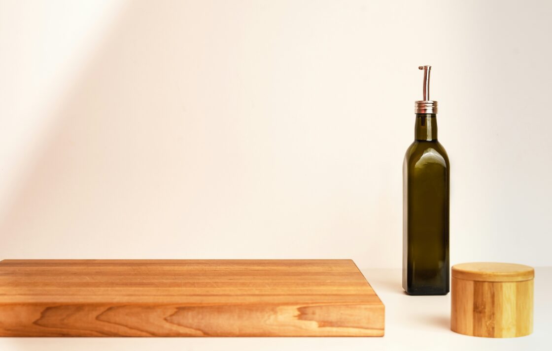 Image of a thick wooden cutting board on a table with a dark glass oil bottle next to it.