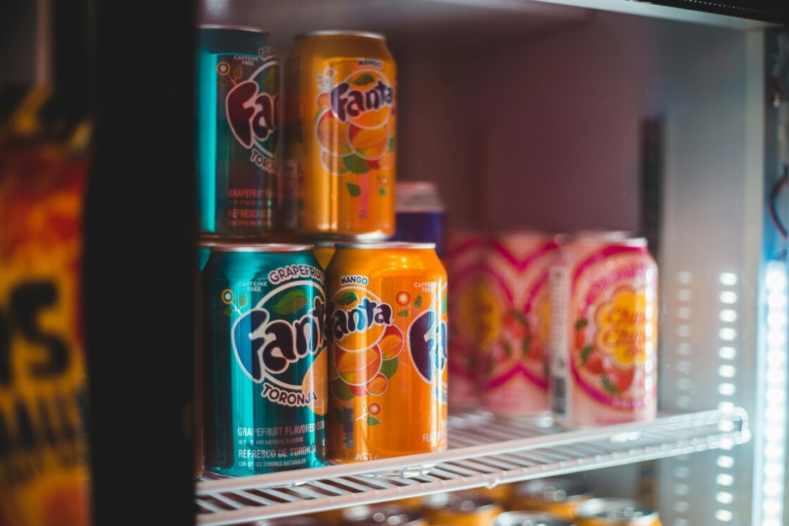 Close-up image of a shelf in a drinks fridge with multiple colorful cans of Fanta and other sugar-sweetened beverages on display.