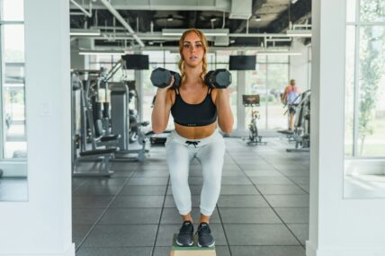 Image of a young woman in a gym, standing on a block in a squat position, with two dumbbells in her hands.